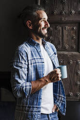 Lächelnder männlicher Fachmann, der wegschaut und eine Kaffeetasse an der Tür hält - EBBF04383