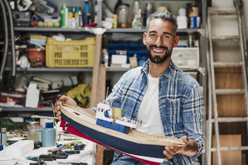 Glücklicher männlicher Handwerker hält ein Schiffsmodell in der Werkstatt - EBBF04388