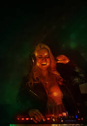 Happy young female DJ in headphones smiling and using synthesizer to play music while standing in smoke during party in nightclub - ADSF28198