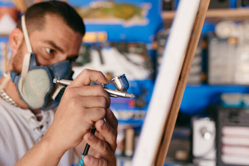 Seitenansicht eines männlichen Künstlers mit Atemschutzmaske, der mit einer Spritzpistole ein Bild auf eine Leinwand malt, während er in einer Kreativwerkstatt arbeitet - ADSF28200