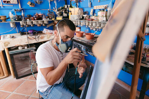 Seitenansicht eines männlichen Künstlers mit Atemschutzmaske, der mit einer Spritzpistole ein Bild auf eine Leinwand malt, während er in einer Kreativwerkstatt arbeitet - ADSF28201