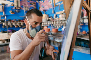 Seitenansicht eines männlichen Künstlers mit Atemschutzmaske, der mit einer Spritzpistole ein Bild auf eine Leinwand malt, während er in einer Kreativwerkstatt arbeitet - ADSF28202