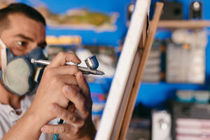 Seitenansicht eines männlichen Künstlers mit Atemschutzmaske, der mit einer Spritzpistole ein Bild auf eine Leinwand malt, während er in einer Kreativwerkstatt arbeitet - ADSF28205