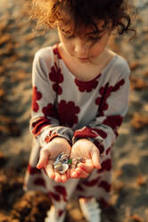 Von oben kleines, lockiges Mädchen im Kleid, das eine Handvoll Muscheln zeigt, während es einen Sommertag am Strand verbringt - ADSF28246