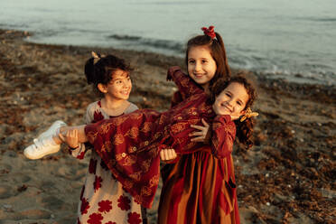 Cute little ethnic girls holding little sister on hands while having fun together on sandy beach near sea - ADSF28249