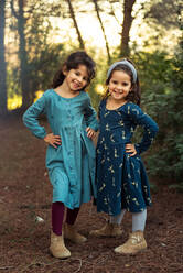 Full body of adorable smiling little sisters in dresses looking at camera while standing together in green forest - ADSF28250