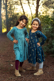 Ganzer Körper von bezaubernden lächelnden kleinen Schwestern in Kleidern, die in die Kamera schauen, während sie zusammen im grünen Wald stehen - ADSF28250