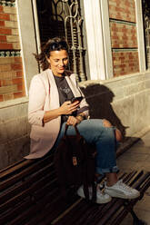 Positive trendy adult ethnic female in ripped jeans and casual blazer sitting on bench near urban building and browsing mobile phone - ADSF28256