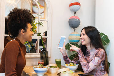 Fröhliche verschiedene weibliche Freunde sitzen am Tisch mit Stöcken in einem Restaurant und benutzen ihr Smartphone, während sie das Wochenende zusammen verbringen - ADSF28265