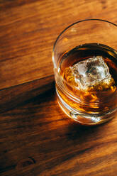 From above glass cup with cold whiskey and cube of ice placed on lumber table in dark room - ADSF28331