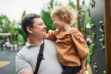 Smiling father embracing cute daughter on playground in summer while looking at each other - ADSF28345