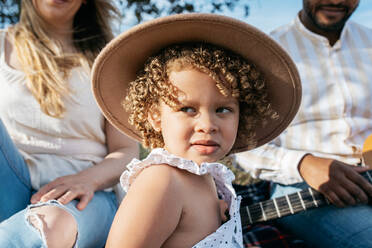 Entzücktes kleines Mädchen mit Hut, das wegschaut, sitzt mit einer gemischtrassigen Familie und genießt ein gemeinsames Picknick, während es in der Natur Gitarre spielt - ADSF28350