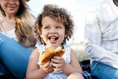 Cheerful little girl eating pastry looking away sitting with multiracial family enjoying picnic together in nature - ADSF28353