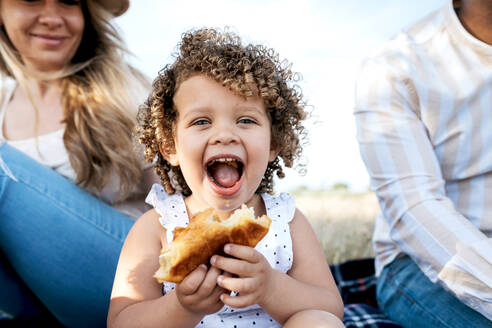 Cheerful little girl eating pastry looking at camera sitting with multiracial family enjoying picnic together in nature - ADSF28354