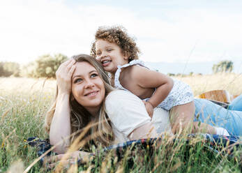 Seitenansicht einer fröhlichen Mutter und eines kleinen Mädchens mit lockigen Haaren, die zusammen auf einer Decke auf einer Wiese liegen und den Sommertag genießen - ADSF28360