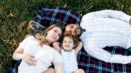 From above of multiethnic family with cute child lying together on blanket on green grass and dreaming - ADSF28363