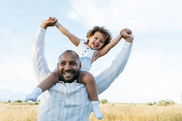 Fröhlicher afroamerikanischer Vater mit süßer kleiner Tochter auf den Schultern, die im Sommer auf einem Feld spielt und Spaß daran hat, wegzuschauen - ADSF28364