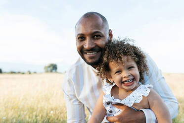African American father holding cute curly haired African American daughter on background of sundown sky in nature looking at camera - ADSF28365