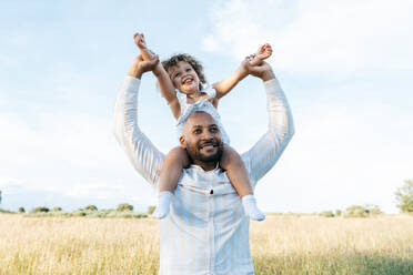 Fröhlicher afroamerikanischer Vater mit süßer kleiner Tochter auf den Schultern, die im Sommer auf einem Feld spielt und Spaß daran hat, wegzuschauen - ADSF28366