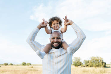 Cheerful African American father with cute little daughter on shoulders playing in field in summer and having fun looking at camera - ADSF28367