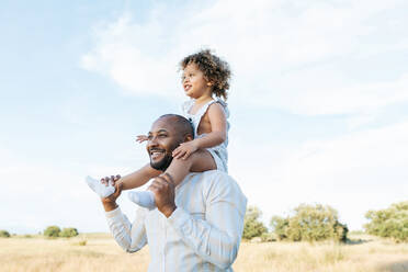 Cheerful African American father with cute little daughter on shoulders playing in field in summer and having fun looking away - ADSF28368