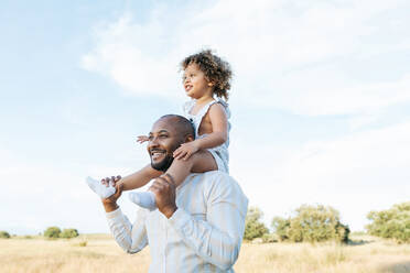 Fröhlicher afroamerikanischer Vater mit süßer kleiner Tochter auf den Schultern, die im Sommer auf einem Feld spielt und Spaß daran hat, wegzuschauen - ADSF28368