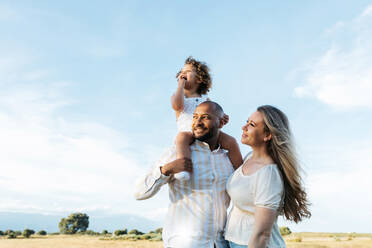 Child sitting on shoulders of father standing in field with mother on summer day - ADSF28373