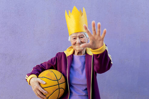 Fröhliche ältere Sportlerin in Sportkleidung mit Basketball, die in die Kamera schaut, während sie eine Kontrollgeste auf lila Hintergrund zeigt - ADSF28384