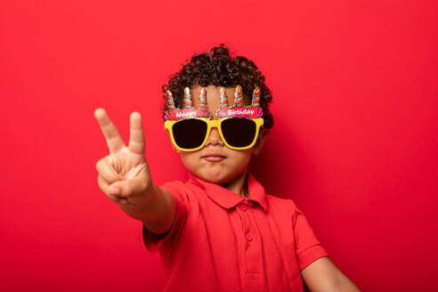 Cooles Kind mit hellen Happy Birthday Sonnenbrille zeigt Frieden Geste auf rotem Hintergrund im Studio - ADSF28390