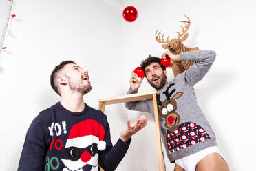 Paar erwachsener schwuler Liebhaber in Unterwäsche und Weihnachtspullis mit dekorativen roten Kugeln, die vor einer weißen Wand mit künstlichem Hirschkopf stehen - ADSF28408