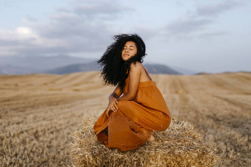 Woman with eyes closed crouching on hay bale during sunset - TCEF02026