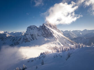 Luftaufnahme des verschneiten Brentenjochs, Österreich, im Winter, mit Kiefern im Vordergrund und Wolken auf dem Gipfel - AAEF12079