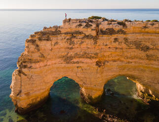 Aerial View of Man standing on the edge of famous cliff formations with arches in Marinha Bay, Portugal with the open atlantic sea in the background - AAEF12090