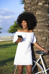 Afro-Frau lacht, während sie mit dem Handy am Fahrrad im Park steht - VEGF04782