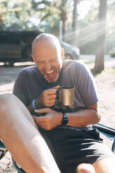 Unbekümmerter Mann mit Kaffeetasse lachend an einem sonnigen Tag im Urlaub - EGHF00131