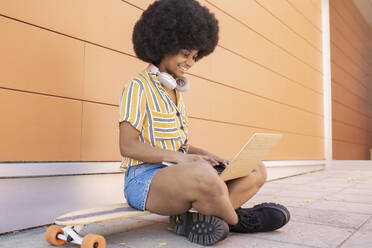 Afro-Frau, die einen Laptop benutzt und im Schneidersitz auf einem Skateboard sitzt - JCCMF03296