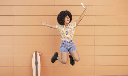 Happy woman taking selfie through smart phone while jumping in front of wall - JCCMF03301