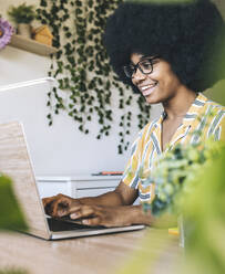 Lächelnde Afro-Frau mit Brille, die bei der Arbeit zu Hause einen Laptop benutzt - JCCMF03308