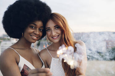 Young female friends smiling while holding sparklers during sunset - JCCMF03355