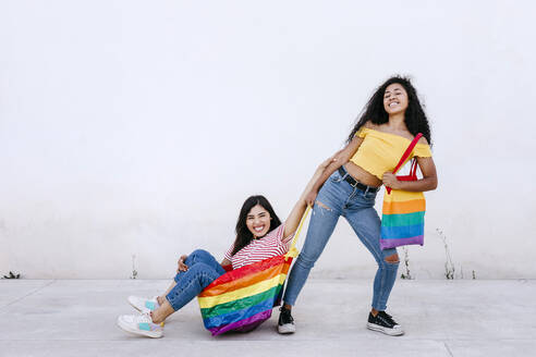 Lächelnde lesbische Frau, die ihre in einer Regenbogentasche sitzende Freundin auf dem Fußweg vor einer weißen Wand zieht - TCEF02046
