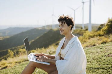 Lächelnde Frau sitzt mit Laptop an einem sonnigen Tag - GMCF00174