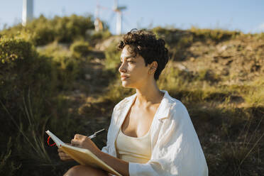 Nachdenkliche Frau sitzt mit Notizbuch an einem sonnigen Tag - GMCF00175