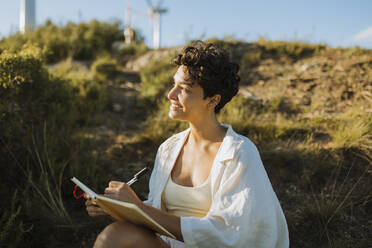 Smiling woman holding pen and notebook while sitting on mountain - GMCF00176