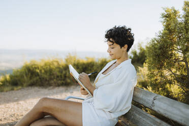 Mid adult woman writing in notebook while sitting on bench at mountain - GMCF00182