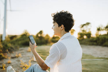 Frau mit kurzen Haaren benutzt Smartphone bei Sonnenuntergang - GMCF00213