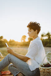 Frau benutzt Mobiltelefon beim Picknick bei Sonnenuntergang - GMCF00215