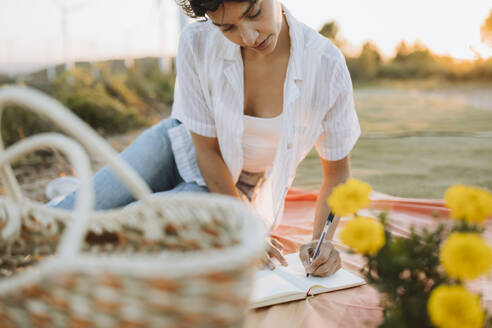 Frau schreibt in einem Buch, während sie bei Sonnenuntergang auf einer Picknickdecke sitzt - GMCF00219