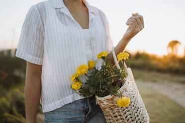 Woman holding bag with yellow flowers during sunset - GMCF00222