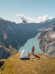 Aerial View of hiker with tent admiring view od Swiss Mountain Lake in Glarus, Switzerland. - AAEF12498