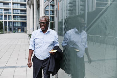Happy businessman with smart phone and blazer walking by reflection on glass wall - ASGF00917